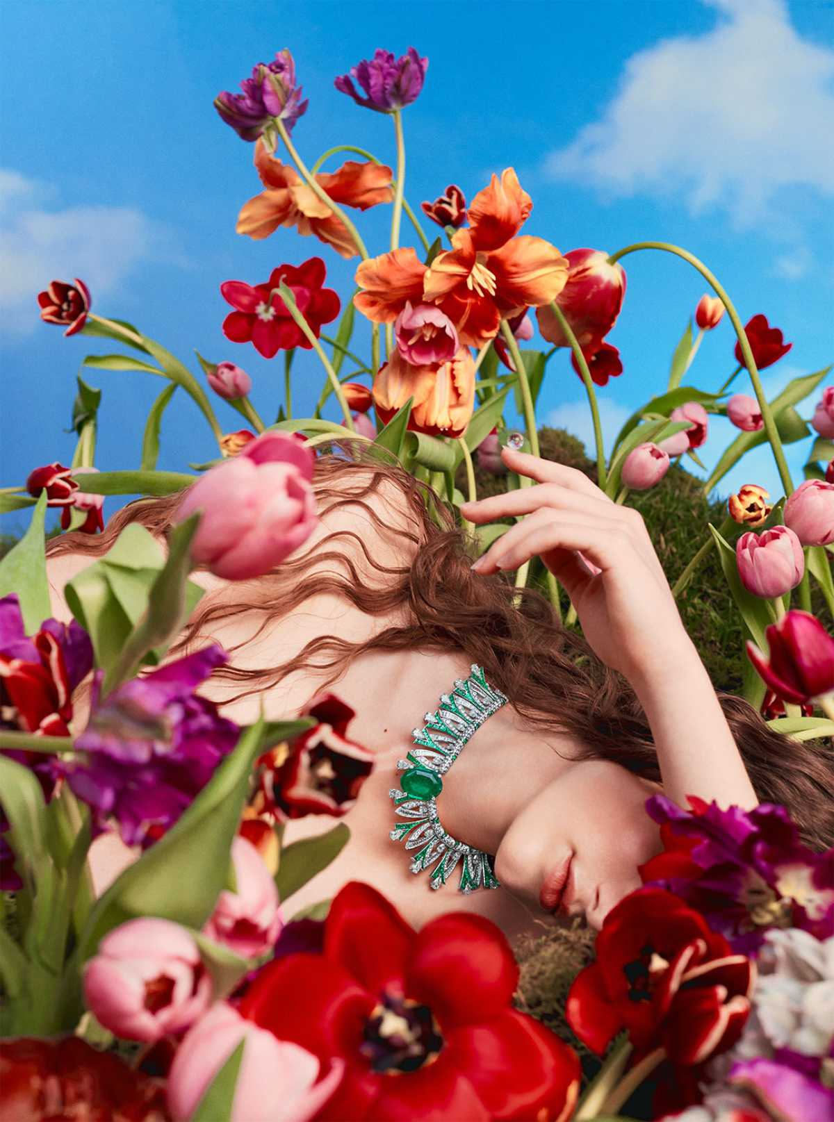 A model reclining among red, coral and pink tulips under a blue sky, wearing a high-jewelry emerald and diamond choker necklace